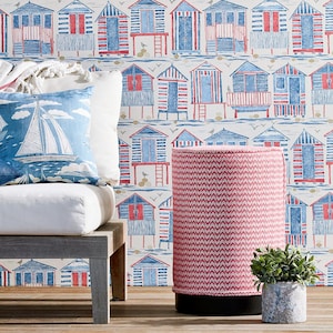 May include: A pink and white chevron patterned cylindrical stool with a black base, in front of a white wall with a blue and red beach hut pattern.