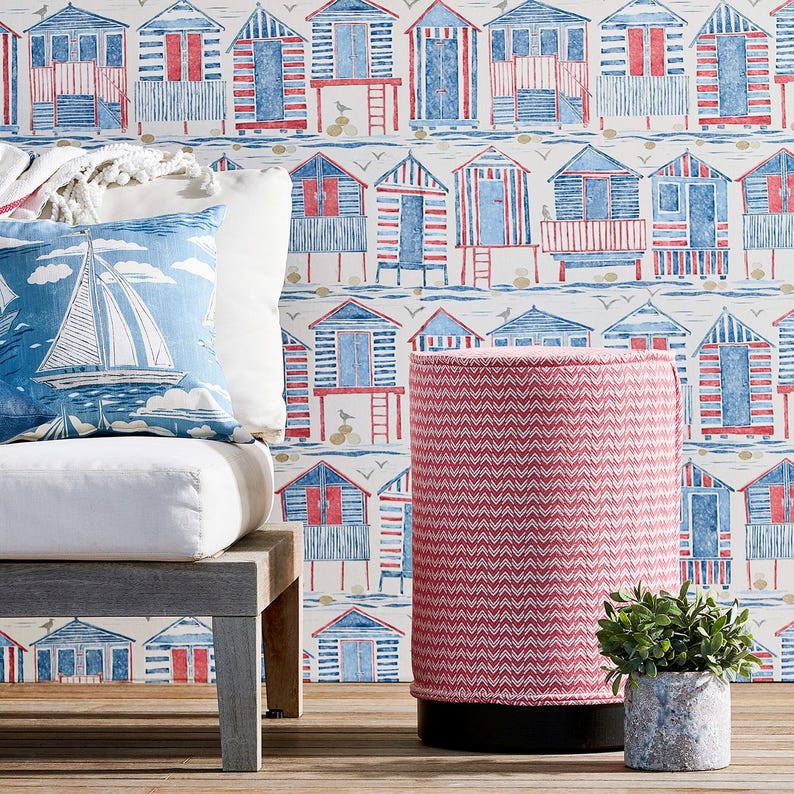 May include: A pink and white chevron patterned cylindrical stool with a black base, in front of a white wall with a blue and red beach hut pattern.