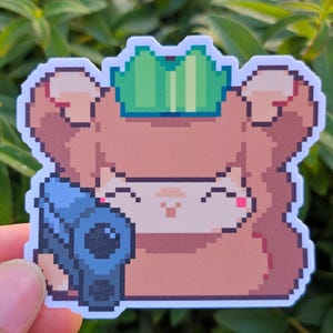 May include: A pixel art sticker of a brown bear wearing a green crown and holding a blue camera. The bear has a happy expression and pink cheeks.