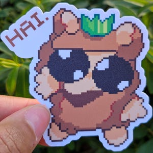 May include: A pixelated brown bear wearing a green crown and sunglasses. The bear is waving with its right paw and has the word "HAI" written above it.