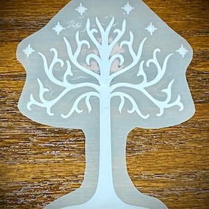 May include: A translucent white tree-shaped design with a detailed tree and star pattern. The design features a central trunk, branches, and roots, with stars at the top. The design is set against a wooden background.