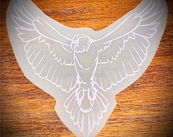 May include: A white, stylized eagle design with outstretched wings. The eagle is depicted in a detailed, line-art style, with feathers and facial features visible. The design is on a light-colored, irregular-shaped background, set against a wooden surface.