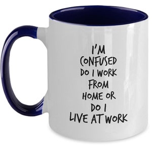 May include: White ceramic coffee mug with a blue rim and handle. The mug has black text that reads "I'm confused do I work from home or do I live at work."