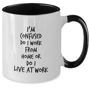 May include: White coffee mug with a black rim and handle. The mug has black text that reads "I'm confused do I work from home or do I live at work."