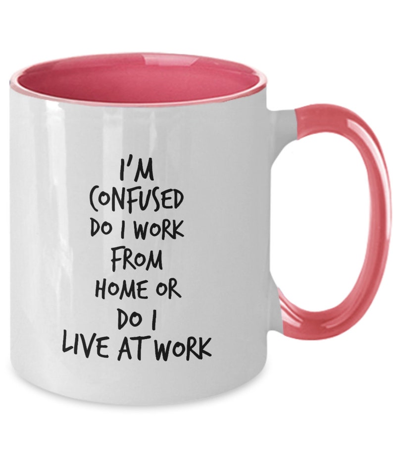 May include: White ceramic coffee mug with a pink rim and handle. The mug has black text that reads "I'm confused do I work from home or do I live at work."