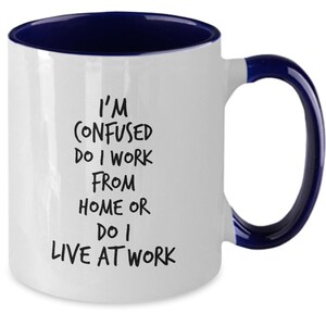 May include: White coffee mug with a blue rim and handle. The mug has black text that reads "I'm confused do I work from home or do I live at work."