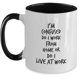 May include: White coffee mug with a black rim and handle. The mug has black text that reads "I'm confused do I work from home or do I live at work."