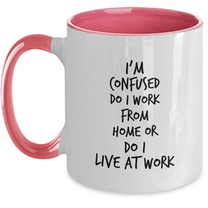 May include: White coffee mug with a pink rim and handle. The mug has black text that reads "I'm confused do I work from home or do I live at work."