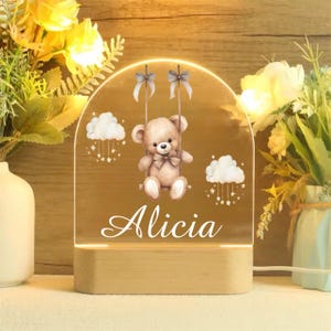 Può includere: Una luce notturna in acrilico bianco con una base in legno. La luce presenta un orsacchiotto di cartone animato su un'altalena con nuvole e stelle. Il nome "Alicia" è scritto sotto l'orso.