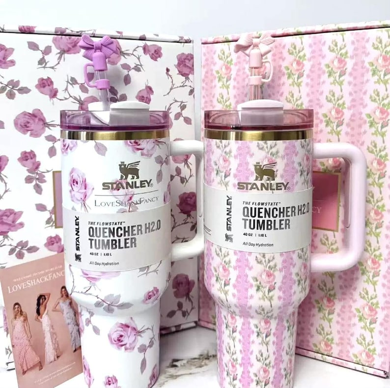 Pu&ograve; includere: Due bicchieri Stanley Quencher H2.0 con motivi floreali e manici coordinati. I bicchieri sono bianchi con rose rosa e finiture dorate. Ogni bicchiere ha una cannuccia ed &egrave; accanto a una scatola con un motivo floreale coordinato. Il testo "LOVESHACKFANCY" &egrave; visibile.
