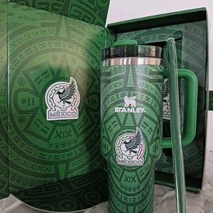 Puede incluir: Una taza de cer&aacute;mica blanca con asa negra y el texto "But First, Coffee" en negrita negra. La taza se encuentra sobre un fondo borroso, sugiriendo un ambiente acogedor.