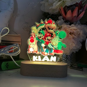May include: A light-up nightlight featuring a colorful image of Mario and friends from the Super Mario Bros. video game series. The nightlight has a wooden base and the name "KIAN" is printed on the front.