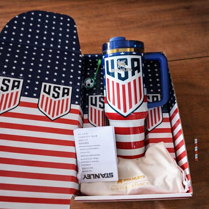 Puede incluir: Un termo de viaje Stanley USA con asa y tapa azules, con un dise&ntilde;o de rayas rojas y blancas con la palabra "USA" en un escudo. El termo est&aacute; empaquetado en una caja con un dise&ntilde;o de bandera americana a juego.