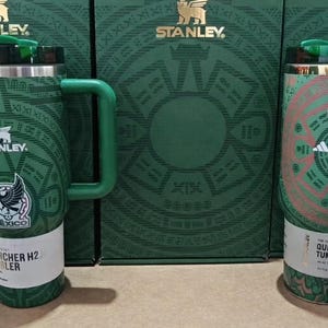 Puede incluir: Vasos Stanley verdes y blancos con el logo del equipo nacional mexicano. Los vasos tienen un asa y una tapa. El fondo presenta cajas verdes con el logo de Stanley. El texto en el vaso dice "Quencher H2.0" y "Quencher Tumbler".
