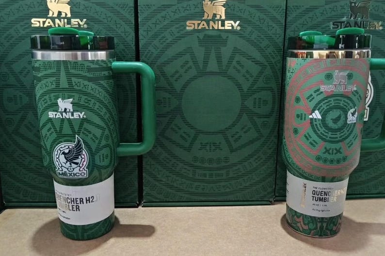 Puede incluir: Vasos Stanley verdes y blancos con el logo del equipo nacional mexicano. Los vasos tienen un asa y una tapa. El fondo presenta cajas verdes con el logo de Stanley. El texto en el vaso dice "Quencher H2.0" y "Quencher Tumbler".