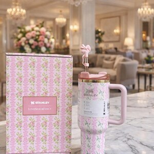 Pu&ograve; includere: Un bicchiere Stanley floreale rosa e bianco con una scatola abbinata. Il bicchiere ha un manico e una cannuccia con un fiore decorativo. La scatola e il bicchiere presentano il logo "LOVESHACKFANCY". Lo sfondo &egrave; un interno lussuoso.