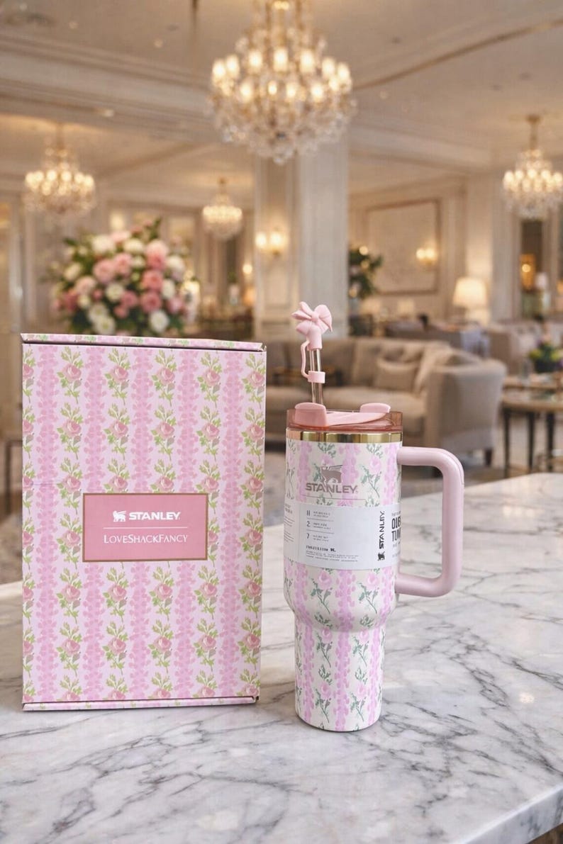 Pu&ograve; includere: Un bicchiere Stanley floreale rosa e bianco con una scatola abbinata. Il bicchiere ha un manico e una cannuccia con un fiore decorativo. La scatola e il bicchiere presentano il logo "LOVESHACKFANCY". Lo sfondo &egrave; un interno lussuoso.