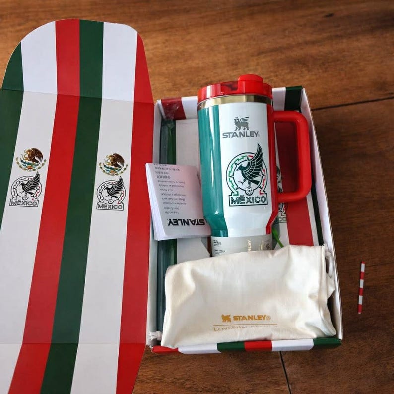 Puede incluir: Un termo de viaje Stanley con asa y tapa rojas, con el escudo de armas mexicano y la palabra "MEXICO". El termo est&aacute; en una caja con los colores de la bandera mexicana y el logotipo de Stanley.