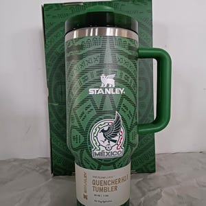 Puede incluir: Vaso Stanley Quencher H2.0 verde con asa, con el emblema nacional mexicano y patrones geom&eacute;tricos. El vaso tiene una tapa negra y una etiqueta blanca con detalles del producto. El embalaje es visible en el fondo.