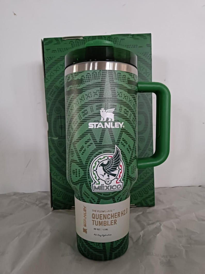 Puede incluir: Vaso Stanley Quencher H2.0 verde con asa, con el emblema nacional mexicano y patrones geom&eacute;tricos. El vaso tiene una tapa negra y una etiqueta blanca con detalles del producto. El embalaje es visible en el fondo.