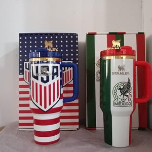 Puede incluir: Dos tazas de viaje aisladas con asas y tapas. Una presenta un dise&ntilde;o de la bandera de EE. UU., la otra un dise&ntilde;o de la bandera mexicana. Ambas tazas tienen el logotipo "Stanley" y se muestran con sus respectivas cajas.