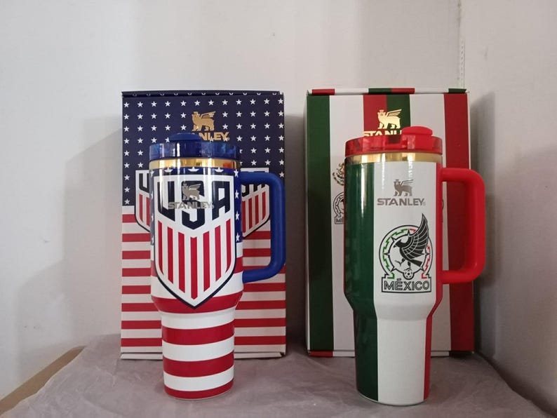 Puede incluir: Dos tazas de viaje aisladas con asas y tapas. Una presenta un dise&ntilde;o de la bandera de EE. UU., la otra un dise&ntilde;o de la bandera mexicana. Ambas tazas tienen el logotipo "Stanley" y se muestran con sus respectivas cajas.
