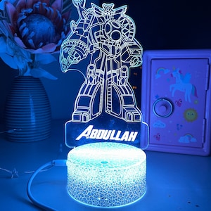 May include: A blue-lit, personalized night light featuring a robot design. The name "ABDULLAH" is illuminated at the base. The light has a cracked-glass effect on the base and is connected to a USB cable. A vase of flowers and a pink safe are in the background.
