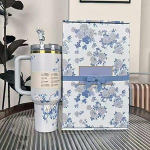 Pu&ograve; includere: Un bicchiere termico bianco con manico e design floreale blu e grigio, accompagnato da una confezione regalo abbinata. Il bicchiere ha una cannuccia e un bordo dorato. La confezione regalo &egrave; decorata con un nastro blu.