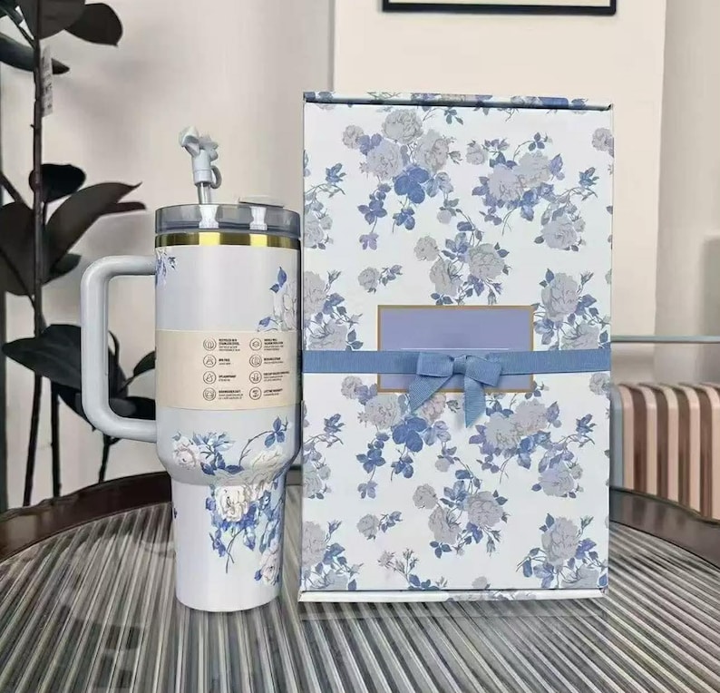 Pu&ograve; includere: Un bicchiere termico bianco con manico e design floreale blu e grigio, accompagnato da una confezione regalo abbinata. Il bicchiere ha una cannuccia e un bordo dorato. La confezione regalo &egrave; decorata con un nastro blu.