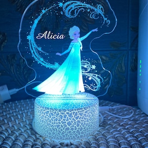 Może przedstawiać: Nocna lampka w kolorze niebieskim i białym z postacią z filmu Kraina Lodu. Postać wyciąga rękę i ma za sobą wirujący wzór. Na lampce widnieje imię "Alicia".