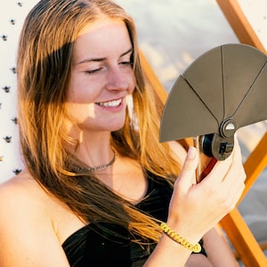 Può includere: Una donna con un top bikini nero e un braccialetto giallo è seduta su una sedia da spiaggia e guarda un supporto per telefono nero con un telefono attaccato.