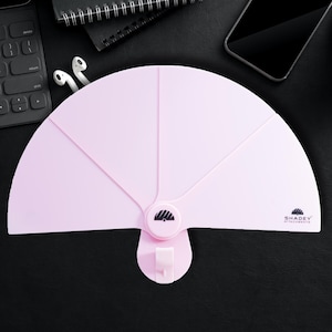 Può includere: Un dispositivo rosa chiaro a forma di ventaglio con un gancio, con il testo "SHADEY ATTACHMENTS". Il dispositivo è progettato per essere fissato a una superficie. La forma a ventaglio ha più sezioni e un punto di attacco centrale.