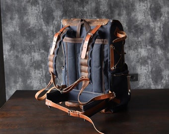 May include: A navy blue backpack with brown leather accents. The backpack features adjustable straps, multiple pockets, and a leather top flap. The bag is made of durable canvas material and is designed for carrying items.