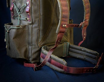 May include: A large, green canvas backpack with brown leather straps and buckles. The backpack has a large main compartment and several smaller pockets.