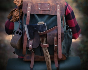 May include: A brown leather backpack with a green canvas body. The backpack has a large axe strapped to the front, along with a leather pouch and other straps. The backpack is being worn by a person in a red and black plaid shirt.