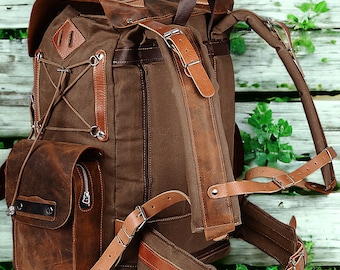 Puede incluir: Una mochila de cuero marrón y lona con correas ajustables y un gran bolsillo frontal. La mochila está sentada sobre un lecho de hierba verde.