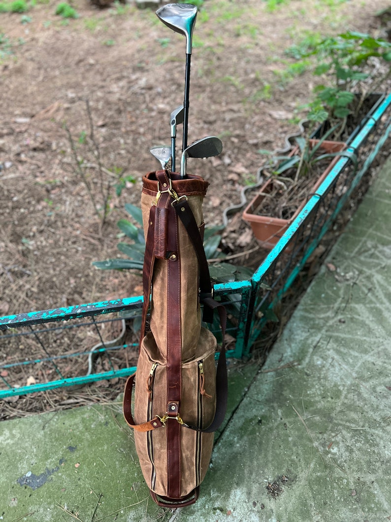 May include: A brown leather and canvas golf bag with a zippered closure and a shoulder strap. The bag is filled with golf clubs.