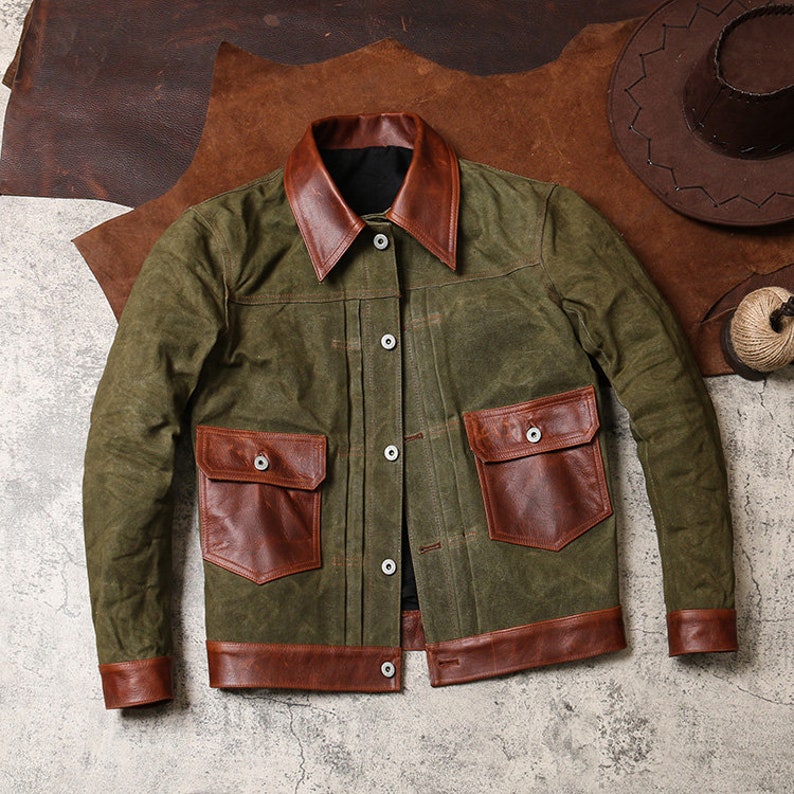 May include: A green canvas jacket with brown leather trim. The jacket has a button-up front, two large pockets on the front, and a collar.