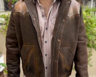 May include: A brown leather jacket with a shearling collar and a zipper closure. The jacket has two flap pockets on the front and a patch pocket on the left chest. The jacket is worn over a plaid shirt and blue jeans.