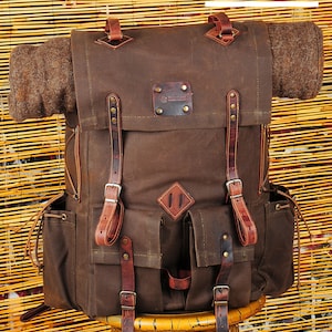 May include: A brown canvas backpack with leather straps and buckles. The backpack has a large main compartment, two front pockets, and a small pocket on the top. The backpack is sitting on a wooden stool.