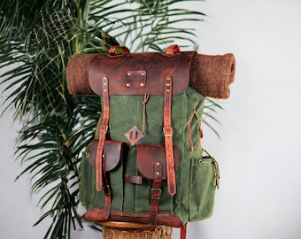 May include: A green canvas backpack with brown leather straps and accents. The backpack has a large main compartment, two smaller front pockets, and a roll-top closure. The backpack is sitting on a wooden stand.