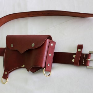 K&ouml;nnte beinhalten: Ein brauner Lederg&uuml;rtel mit einer passenden braunen Ledertasche. Die Tasche hat zwei Metallringe und eine Klappe mit einem dekorativen N&auml;hten-Detail. Der G&uuml;rtel hat eine silberne Schnalle.