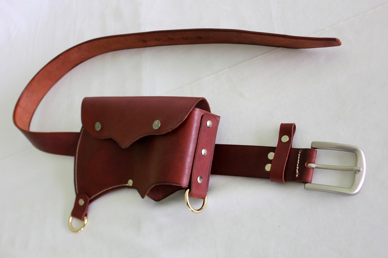K&ouml;nnte beinhalten: Ein brauner Lederg&uuml;rtel mit einer passenden braunen Ledertasche. Die Tasche hat zwei Metallringe und eine Klappe mit einem dekorativen N&auml;hten-Detail. Der G&uuml;rtel hat eine silberne Schnalle.