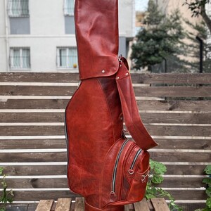 Puede incluir: Una bolsa de golf de cuero marr&oacute;n rojizo con una correa para el hombro y m&uacute;ltiples compartimentos con cremallera. La bolsa tiene una forma alta y cil&iacute;ndrica con una base redondeada y est&aacute; dise&ntilde;ada para guardar palos de golf. La bolsa est&aacute; sobre una estructura de madera.