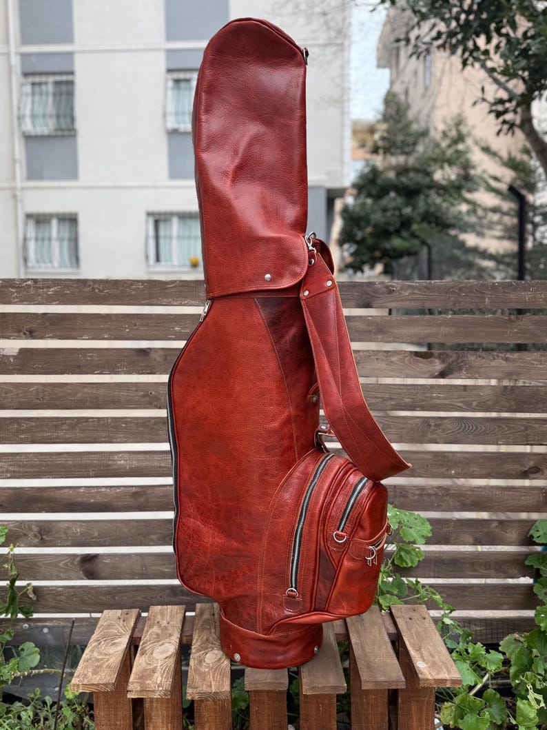 Puede incluir: Una bolsa de golf de cuero marr&oacute;n rojizo con una correa para el hombro y m&uacute;ltiples compartimentos con cremallera. La bolsa tiene una forma alta y cil&iacute;ndrica con una base redondeada y est&aacute; dise&ntilde;ada para guardar palos de golf. La bolsa est&aacute; sobre una estructura de madera.
