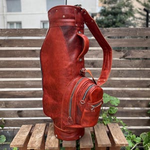 Puede incluir: Una bolsa de golf de cuero marr&oacute;n rojizo con una correa para el hombro y m&uacute;ltiples bolsillos con cremallera. Un palo de golf est&aacute; insertado en la parte superior de la bolsa. La bolsa est&aacute; sobre un palet de madera.