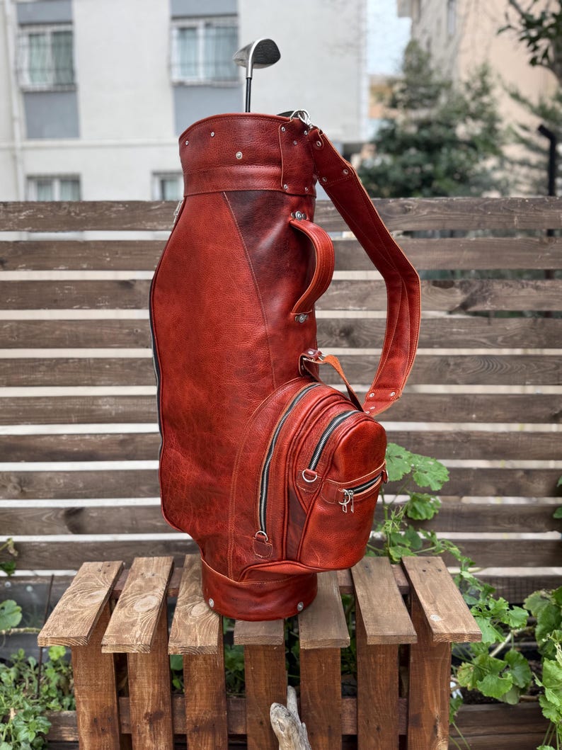Puede incluir: Una bolsa de golf de cuero marr&oacute;n rojizo con una correa para el hombro y m&uacute;ltiples bolsillos con cremallera. Un palo de golf est&aacute; insertado en la parte superior de la bolsa. La bolsa est&aacute; sobre un palet de madera.