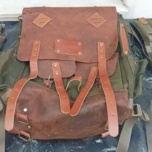 May include: A brown leather backpack with green canvas accents and multiple straps. The backpack has a large flap with a leather pocket and two diamond-shaped leather accents.