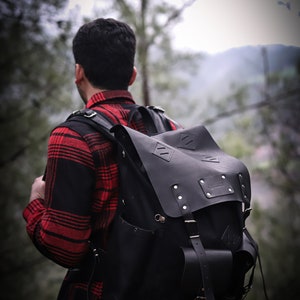 May include: A person wearing a red and black plaid shirt with a black leather backpack. The backpack has a large flap with a buckle and a smaller pocket on the front.