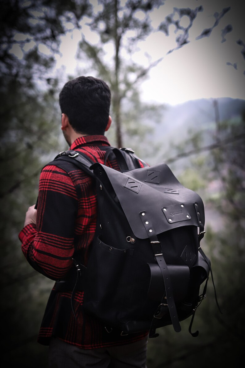 May include: A person wearing a red and black plaid shirt with a black leather backpack. The backpack has a large flap with a buckle and a smaller pocket on the front.